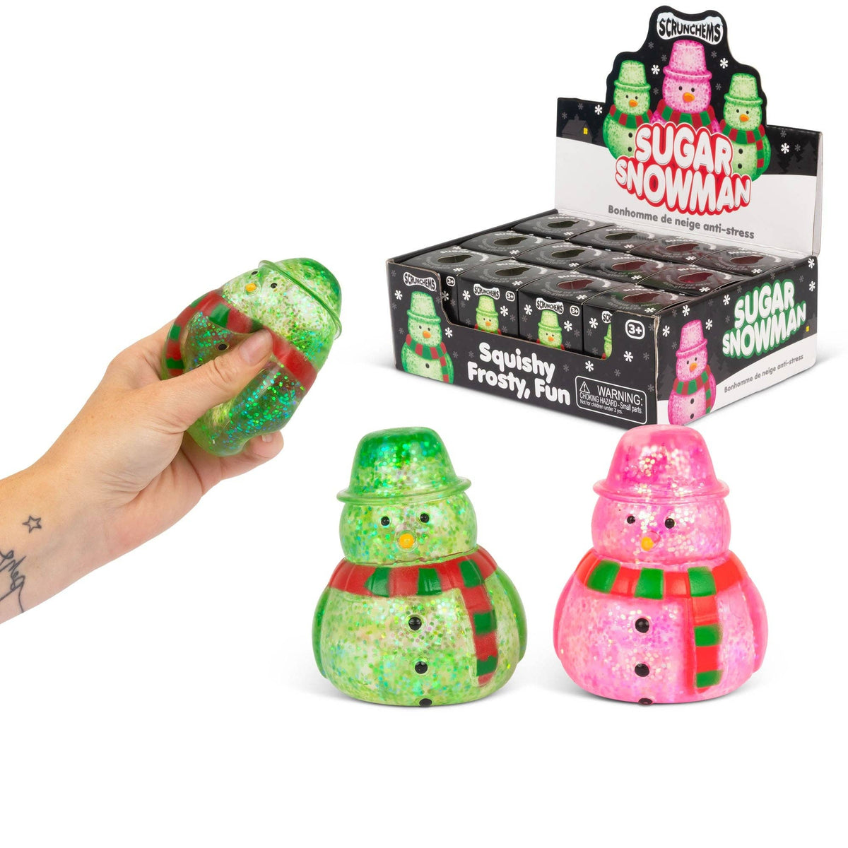 A hand holds a green glittery squishy snowman toy, with two more snowman toys in front and a display box labeled 'Sugar Snowman' in the background. The toys are green and pink glittery snowmen with red and green striped scarves.