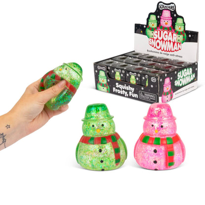 A hand holds a green glittery squishy snowman toy, with two more snowman toys in front and a display box labeled 'Sugar Snowman' in the background. The toys are green and pink glittery snowmen with red and green striped scarves.
