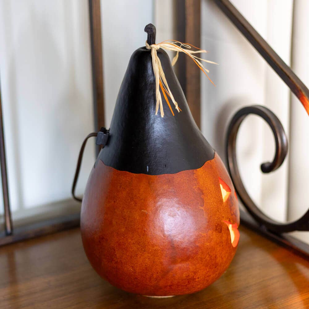 A decorative carved gourd pumpkin with a black top and a warm orange-brown body, lit from within, sitting on a wooden surface.