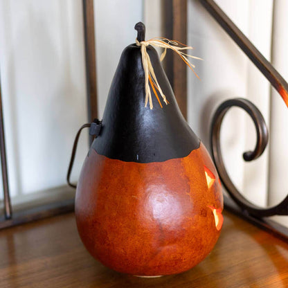 A decorative carved gourd pumpkin with a black top and a warm orange-brown body, lit from within, sitting on a wooden surface.