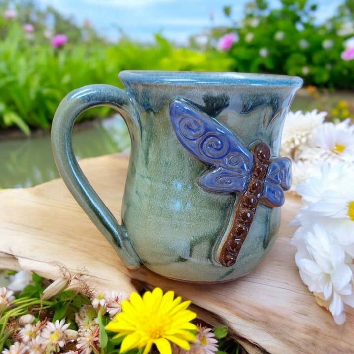 Ceramic mug with dragonfly design on a wooden surface with flowers in the background