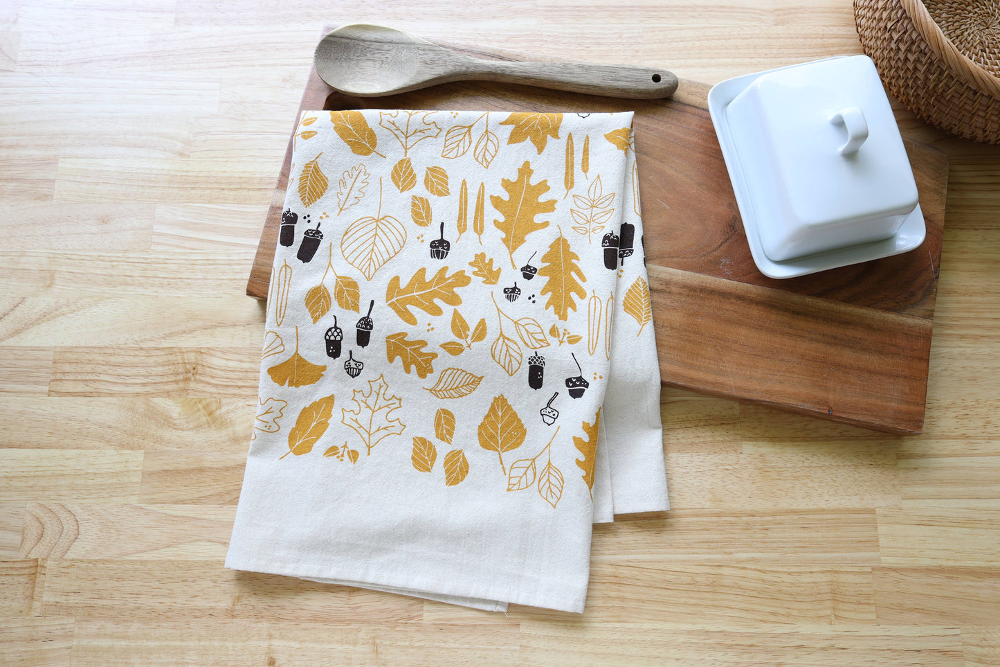 A folded white kitchen towel with a mustard yellow and dark brown autumn leaf pattern, styled on a wooden cutting board with a wooden spoon and a white butter dish on a light wood tabletop.