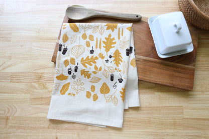 A folded white kitchen towel with a mustard yellow and dark brown autumn leaf pattern, styled on a wooden cutting board with a wooden spoon and a white butter dish on a light wood tabletop.