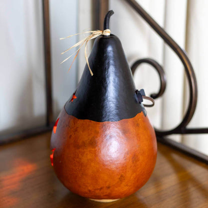 A decorative carved gourd lantern with a glossy orange-brown lower half and a black upper half, topped with a small dried stem and tied with twine, shown on a wooden surface.