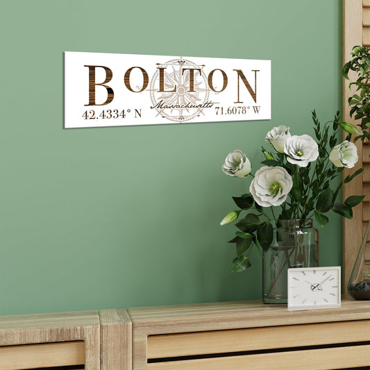 Decorative sign with 'Bolton, Massachusetts' and coordinates on a green wall above a wooden surface with flowers and a clock.