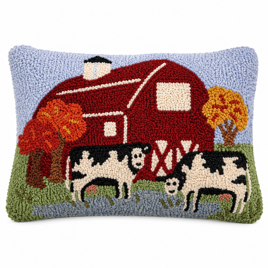 A rectangular hand-hooked wool pillow featuring a farm scene with a red barn, black-and-white cows, green grass, and autumn trees on a light blue background.