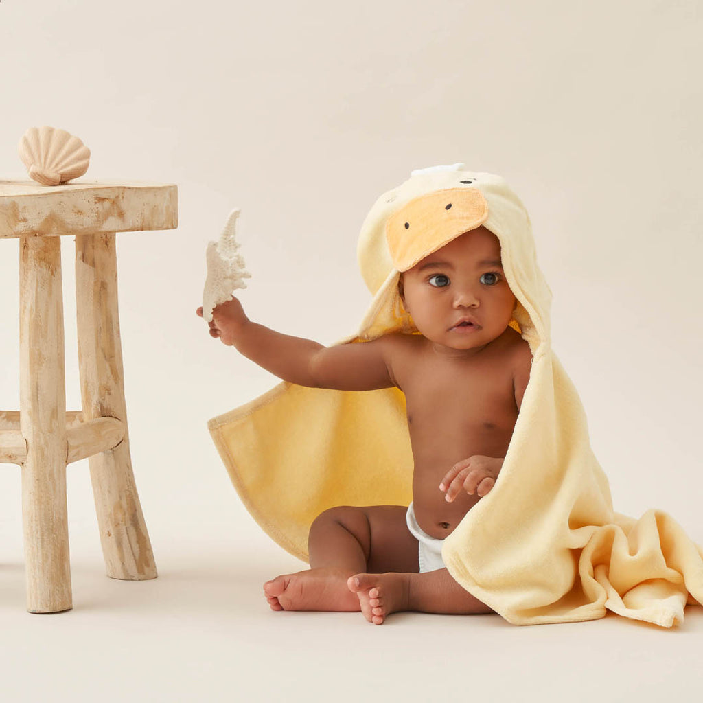 Baby wrapped in a yellow towel with duck design, holding a shell, on a light background