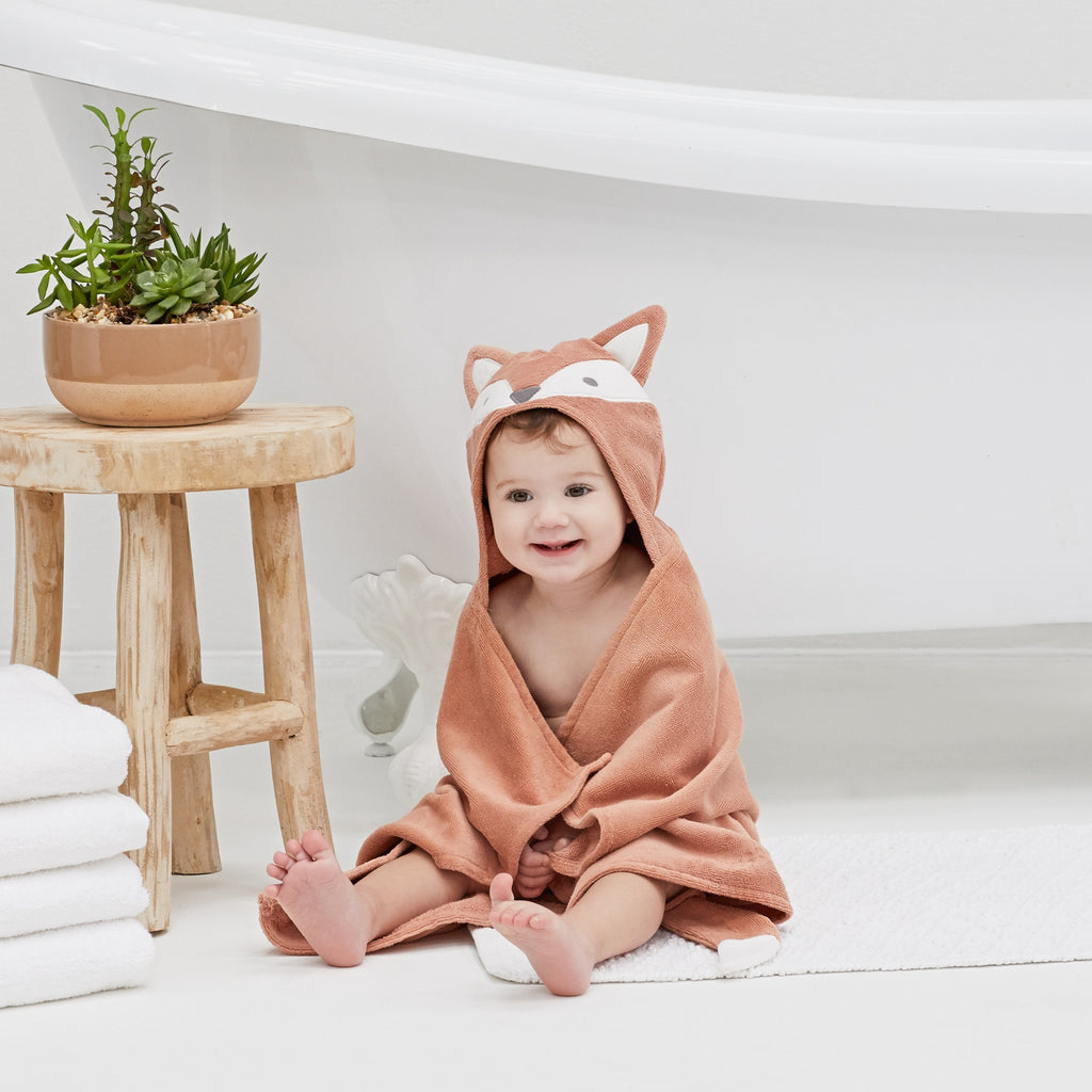 Baby wrapped in a fox-themed hooded towel sitting on a white surface with a wooden stool and plant in the background.