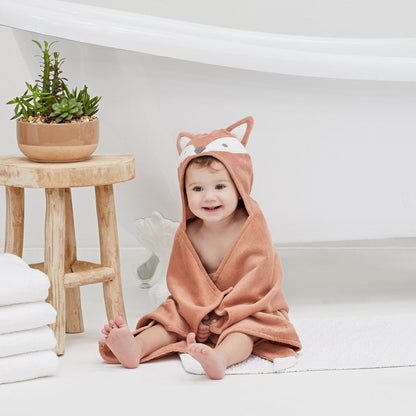 Baby wrapped in a fox-themed hooded towel sitting on a white surface with a wooden stool and plant in the background.