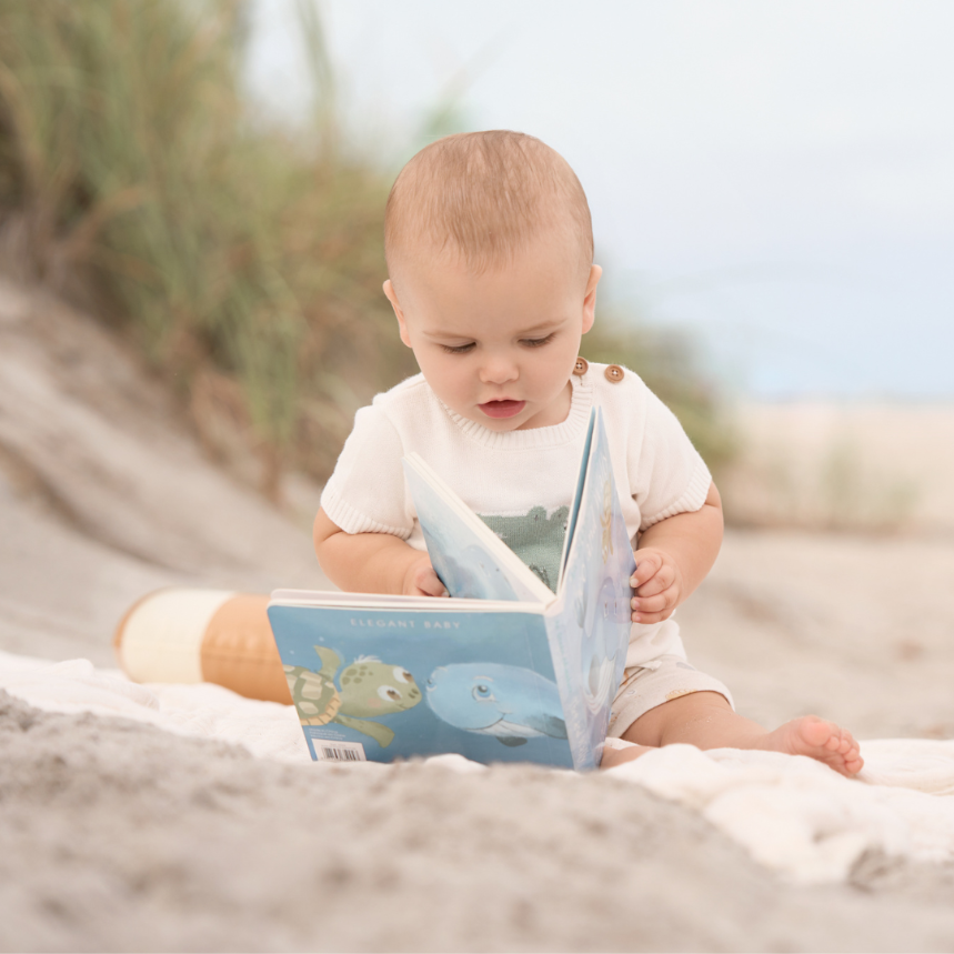Baby sitting on sand reading a book with beach background