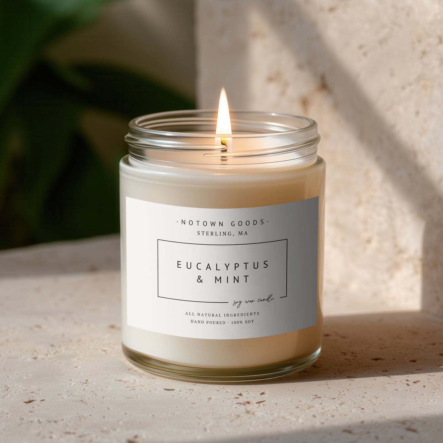Clear jar candle with white wax and a decorative label on a blurred outdoor background.