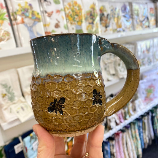 Handmade ceramic mug with honeycomb design and bee decorations held in front of a store shelf.