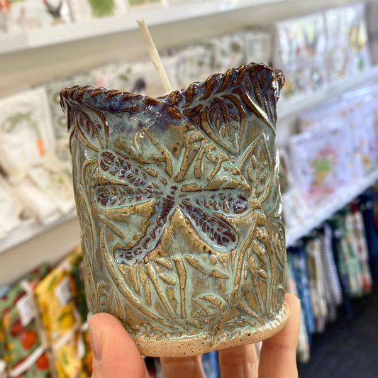 Handmade ceramic candle with leaf and dragonfly design held in front of a store shelf.