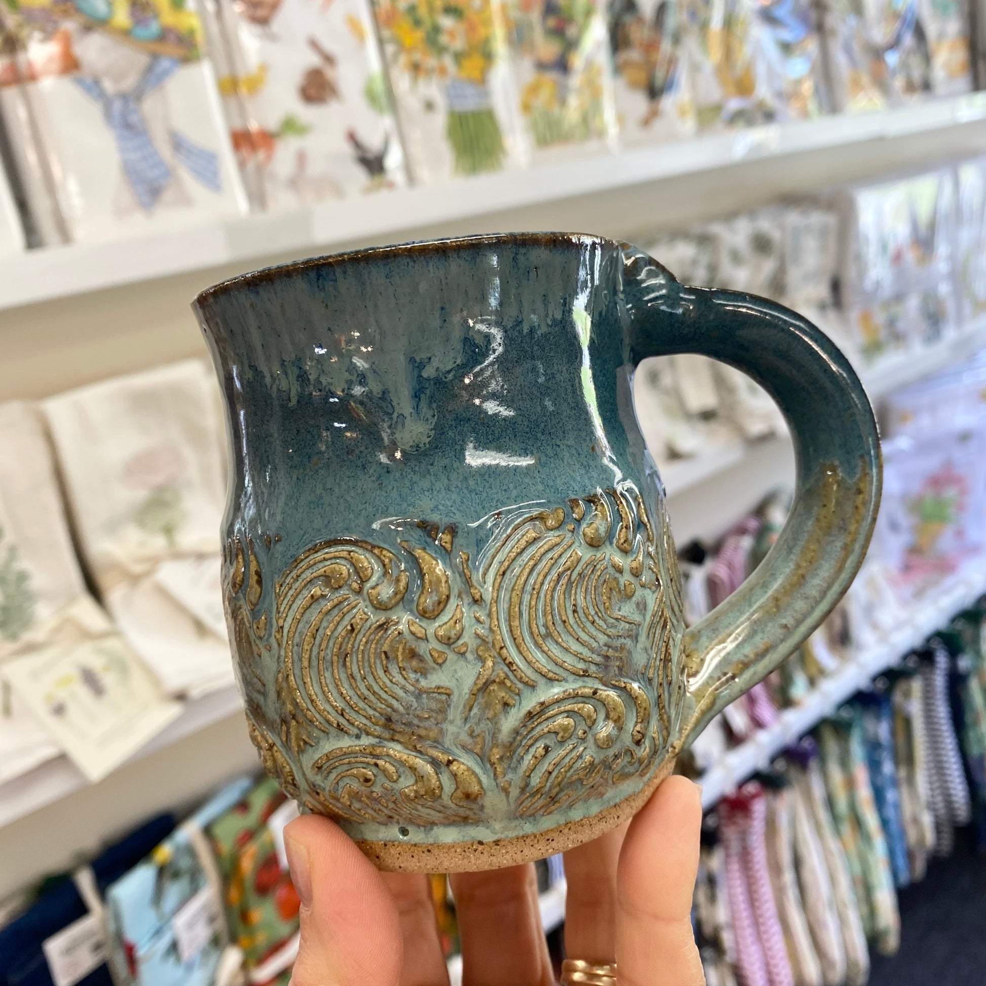 Hand holding a blue ceramic mug with an ocean wave pattern in a store setting.
