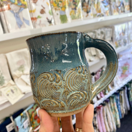 Hand holding a blue ceramic mug with an ocean wave pattern in a store setting.