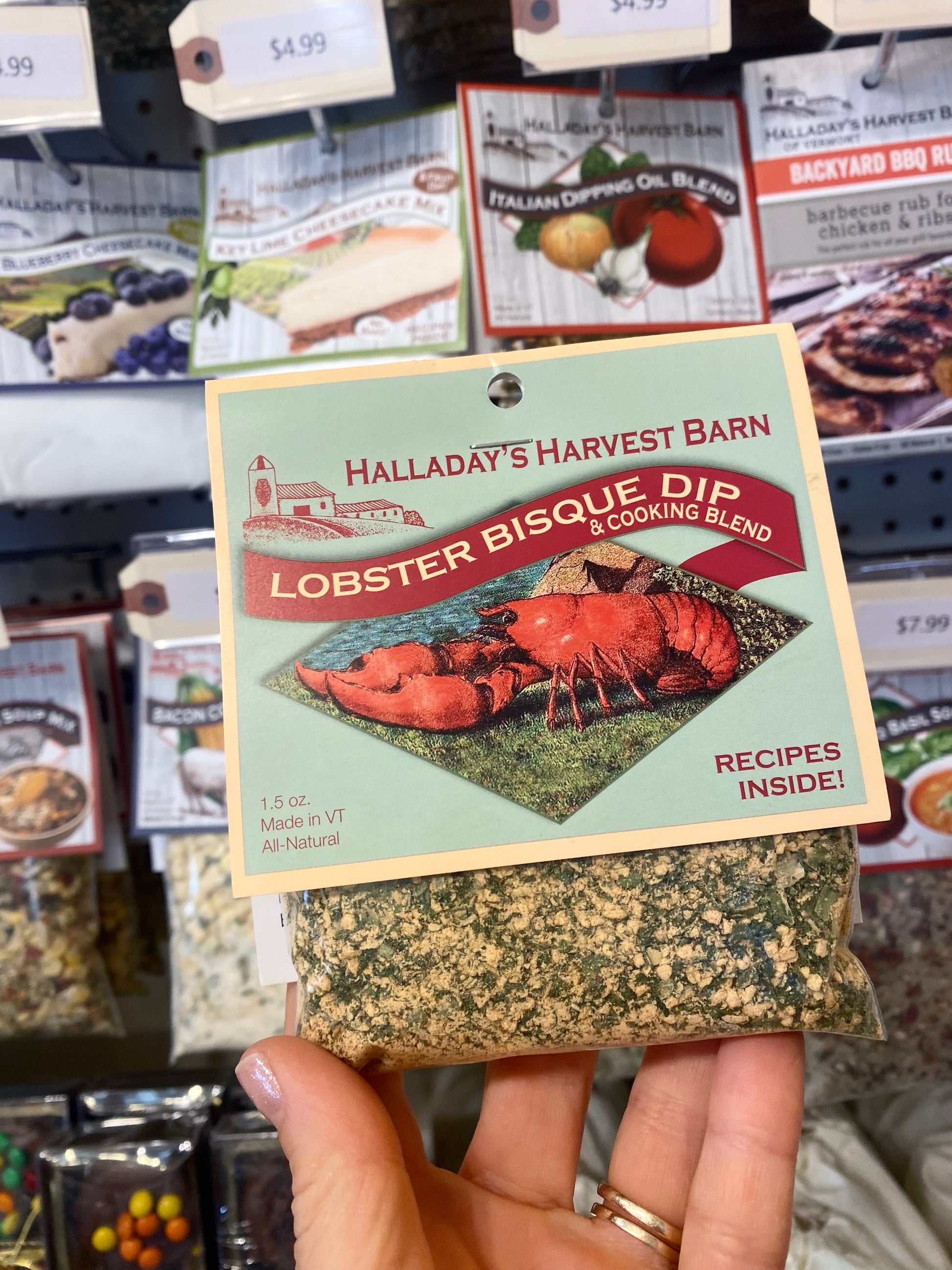 Hand holding a package of Halladay's Harvest Barn Lobster Bisque Dip with a store shelf in the background.