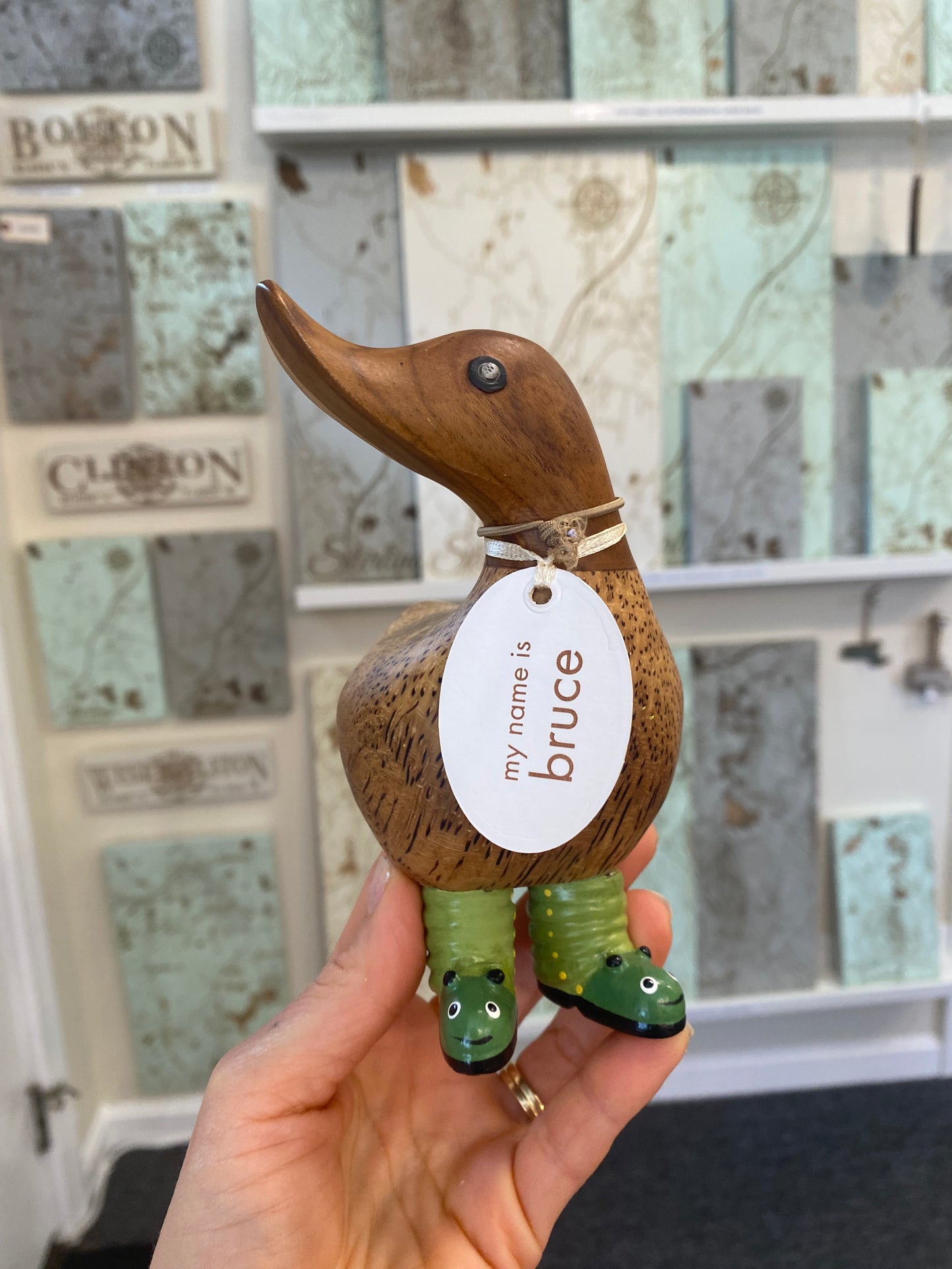 Wooden duck figurine with a tag and green boots, held by a hand in front of a decorative wall.