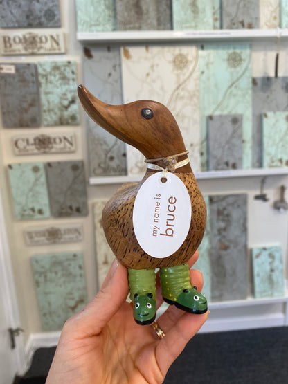 Wooden duck figurine with a tag and green boots, held by a hand in front of a decorative wall.