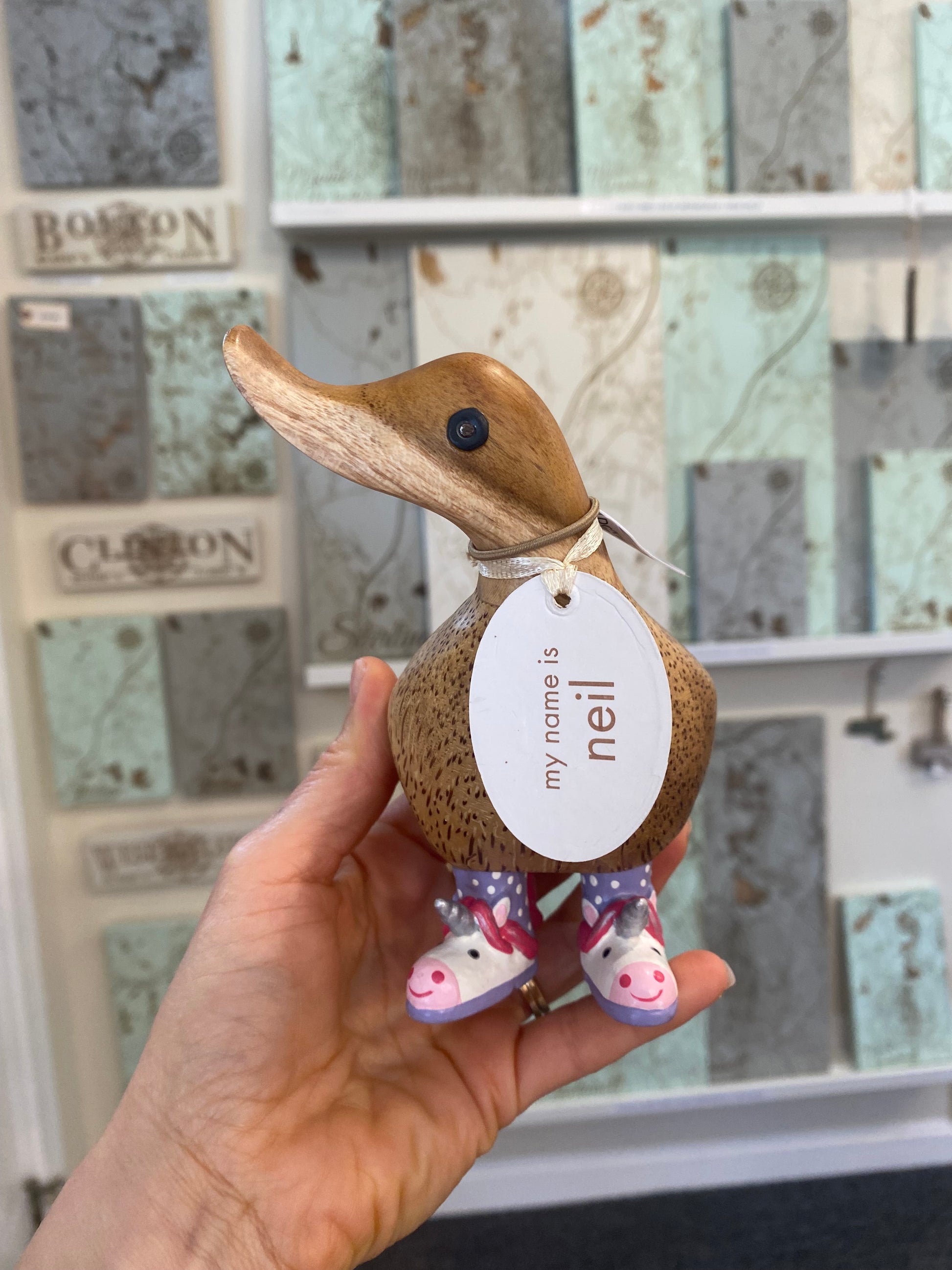 Wooden duck figurine with a name tag held by a hand in front of a decorative wall.