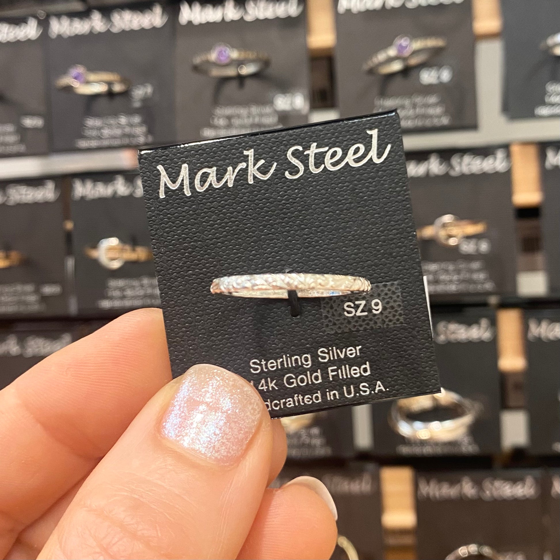 Hand holding a Mark Steel jewelry card with a silver ring attached, against a background of similar cards.
