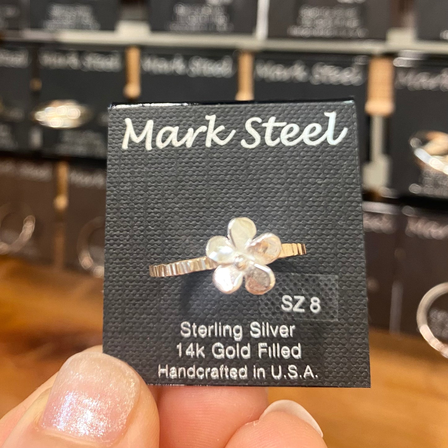 Flower-shaped ring with 'Mark Steel' packaging in a store setting