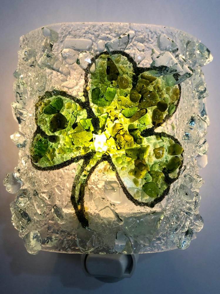 A square glass night light featuring a green shamrock design embedded in clear, textured broken glass, glowing with a warm light from within.