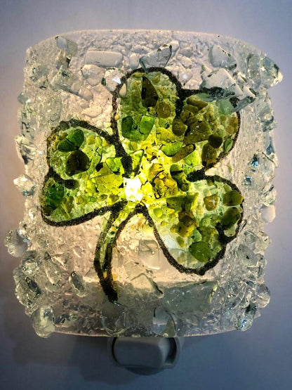A square glass night light featuring a green shamrock design embedded in clear, textured broken glass, glowing with a warm light from within.