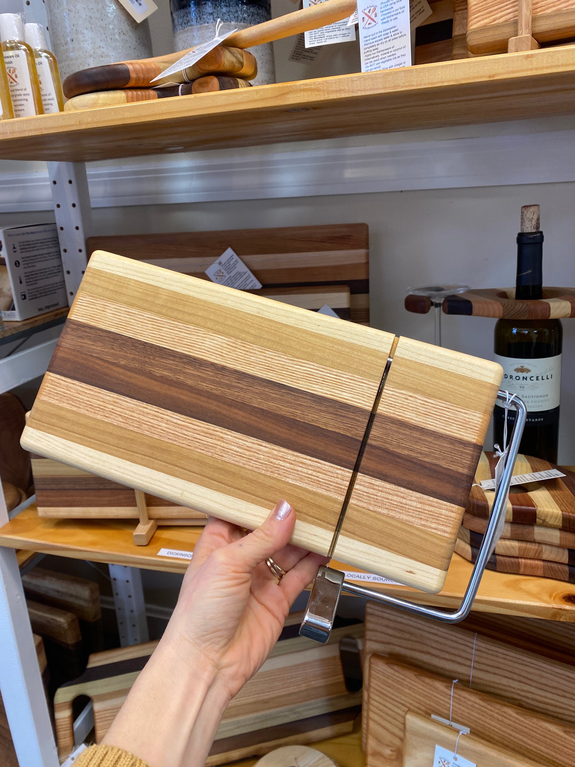 Hand holding a wooden cutting board with a cheese slicing wire in a store setting.