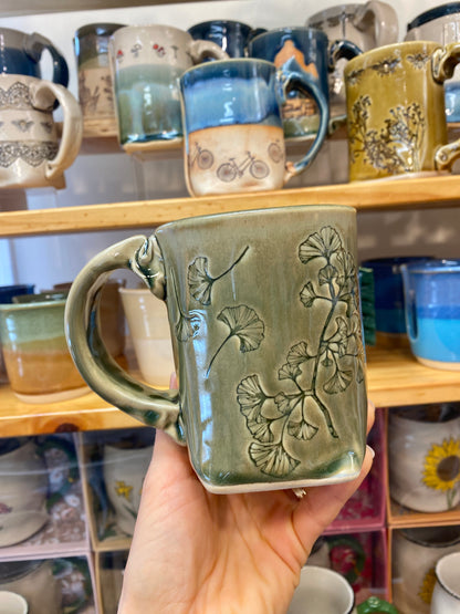 Hand holding a green ceramic mug with ginko leaves pattern against a background of shelves with various mugs.