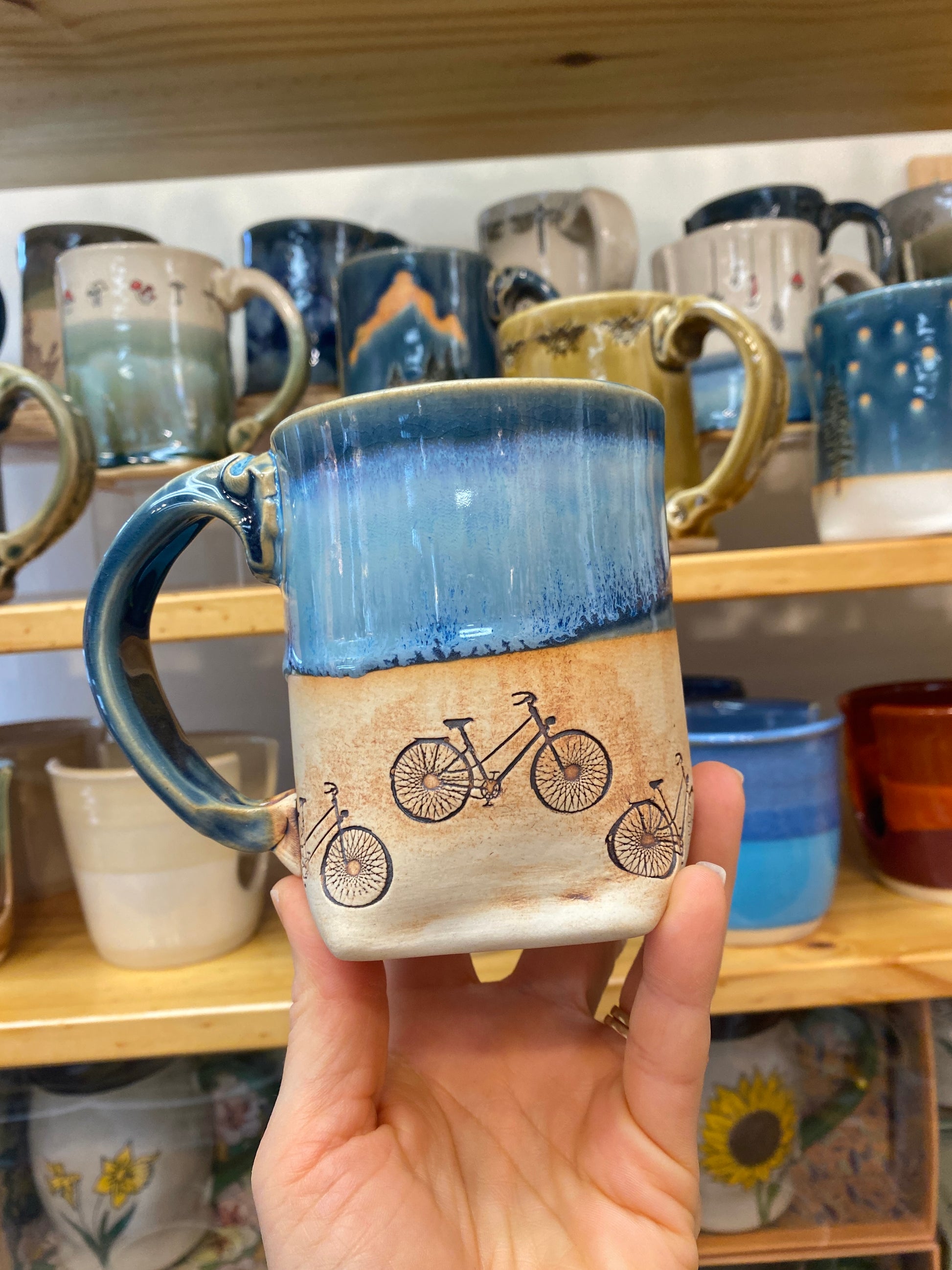 Hand holding a ceramic mug with blue top and handle and bicycle design on beige exterior at the bottom, against a background of shelves with various mugs.