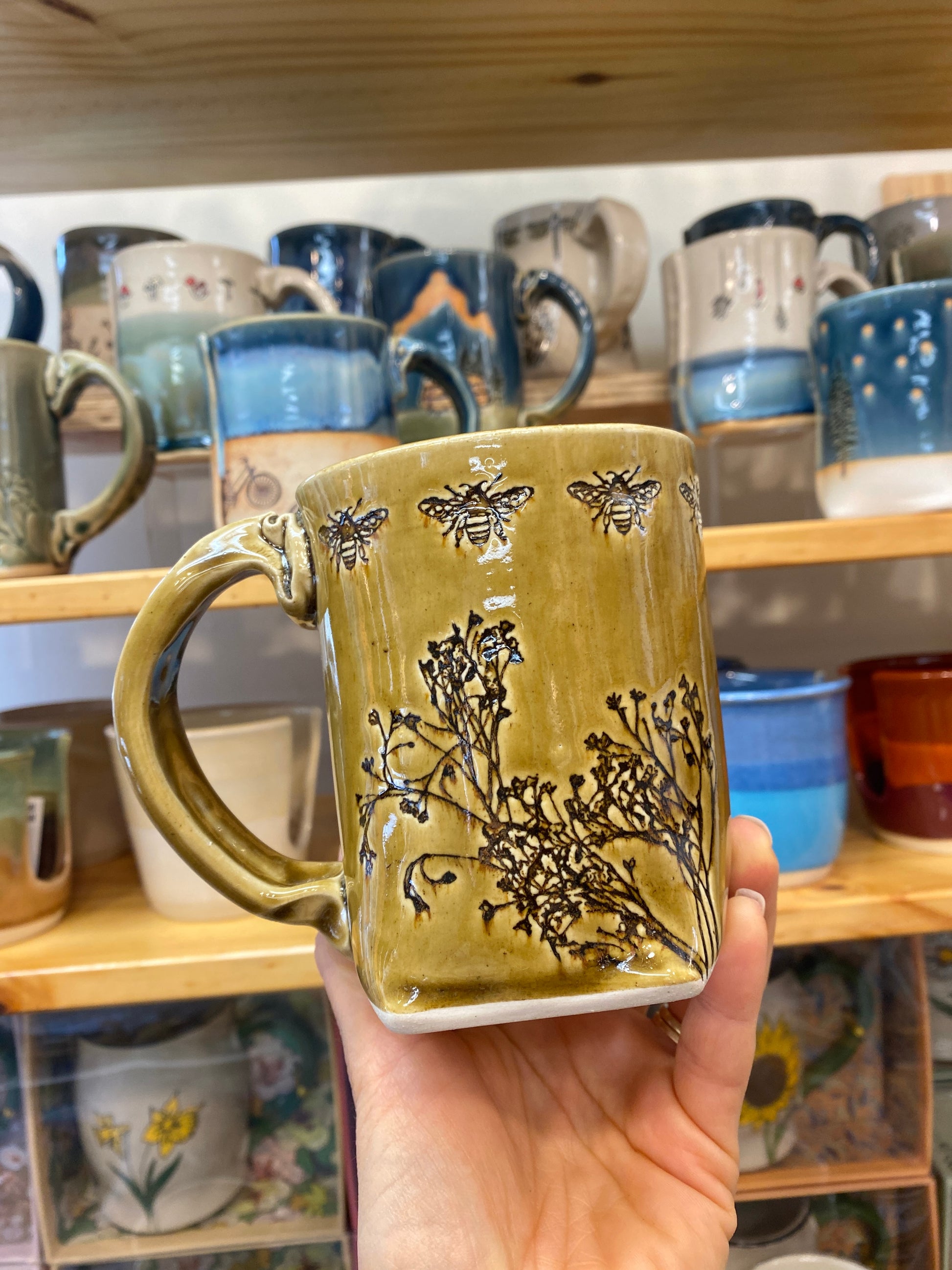 Hand holding a golden brown ceramic mug with bee and floral design in front of a shelf of other mugs.