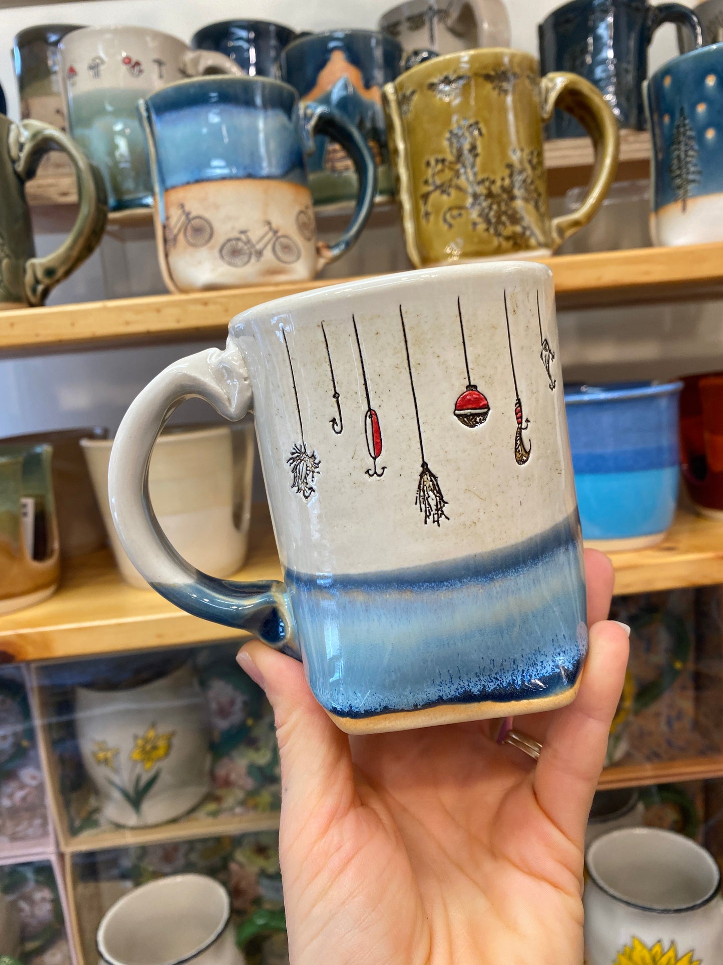 Hand holding a ceramic mug with fishing-themed design in front of a shelf with various mugs.