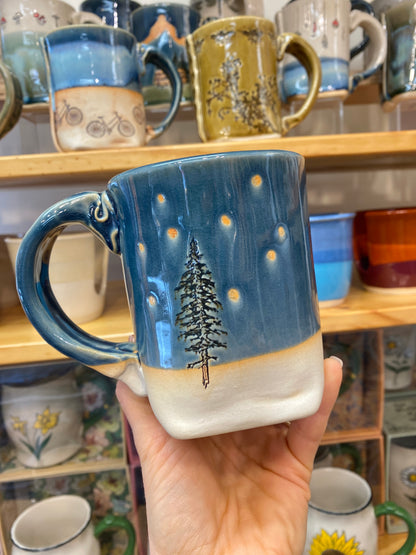 Hand holding a blue mug with a tree design against a background of shelves with various mugs.