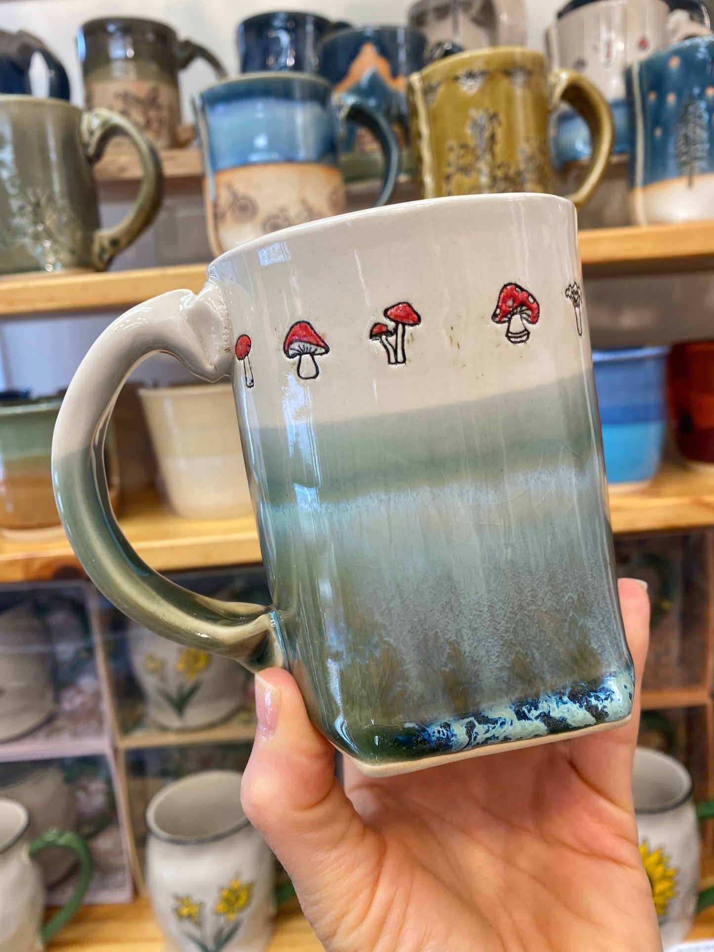 Hand holding a ceramic mug with mushroom design in front of a shelf of mugs.