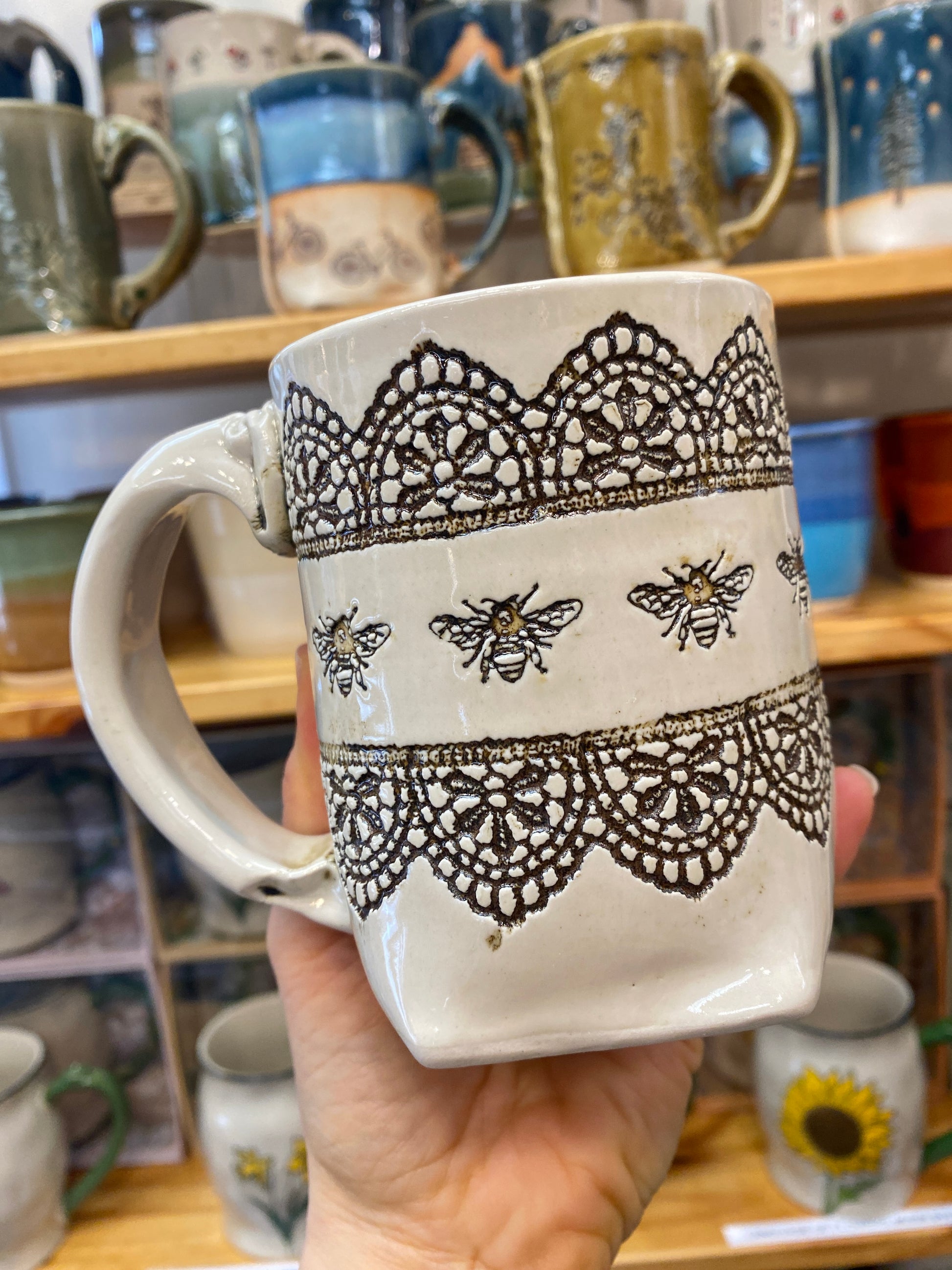 White mug with black lace and bee design held in front of a shelf with various mugs.
