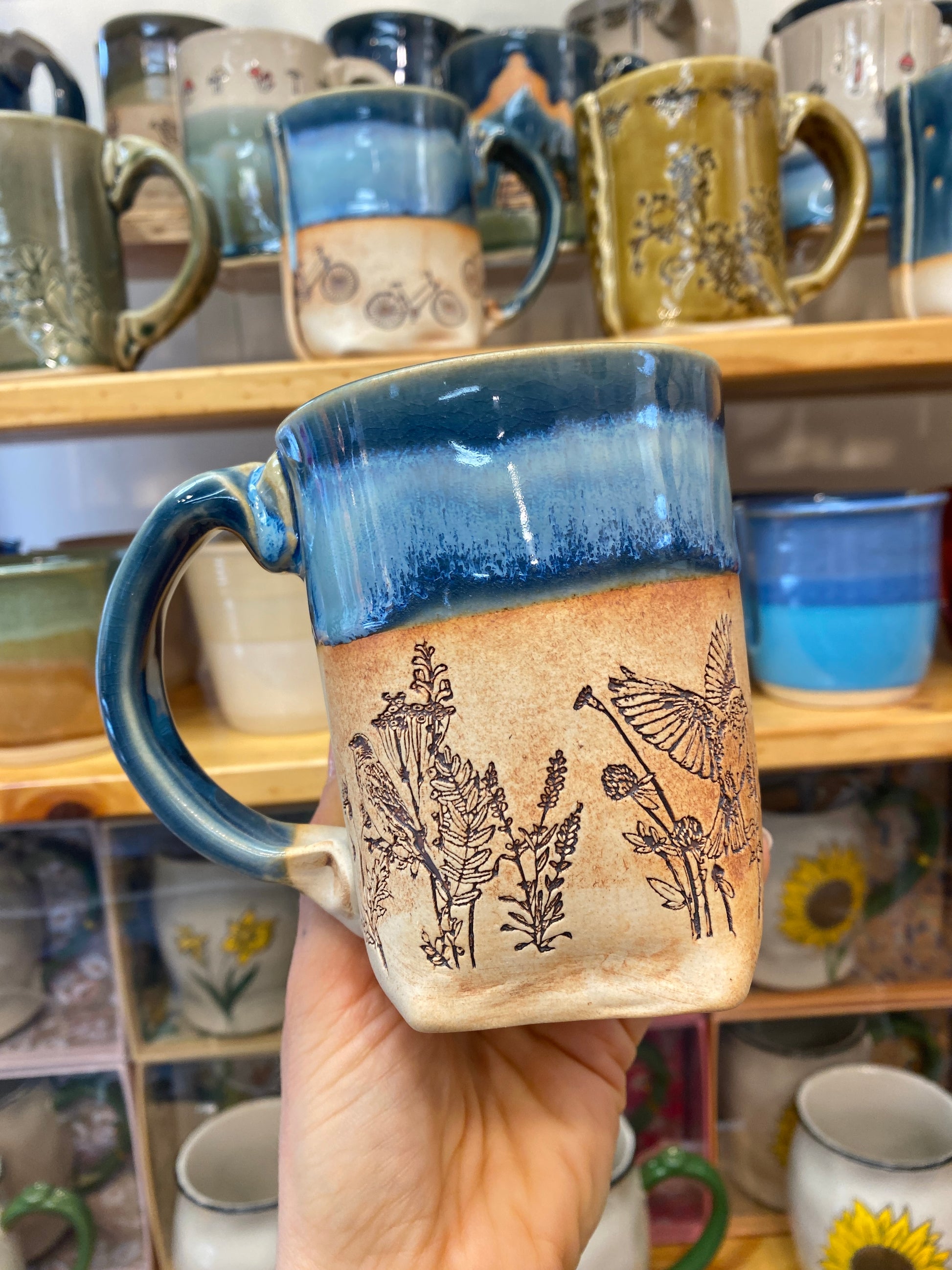 Handmade mug with floral design held in front of a display of other mugs.