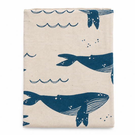 A cream colored tea towel with blue whale and waves design on a white background.