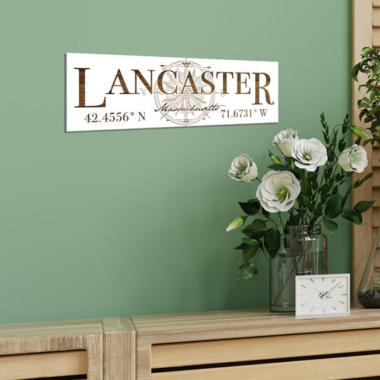 Decorative sign with 'Lancaster, Massachusetts' and coordinates on a green wall above a wooden surface with flowers and a clock.