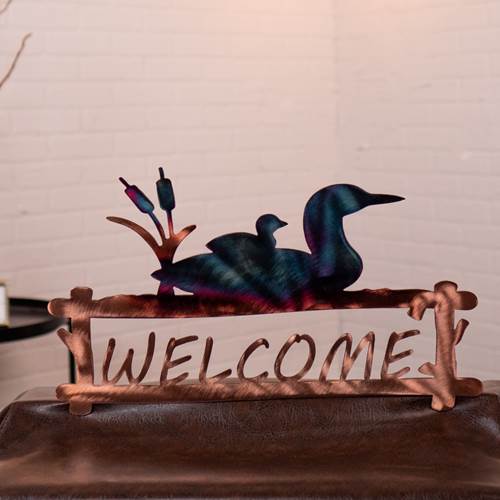 Decorative metal sign with loons and 'WELCOME' text on a wooden surface.