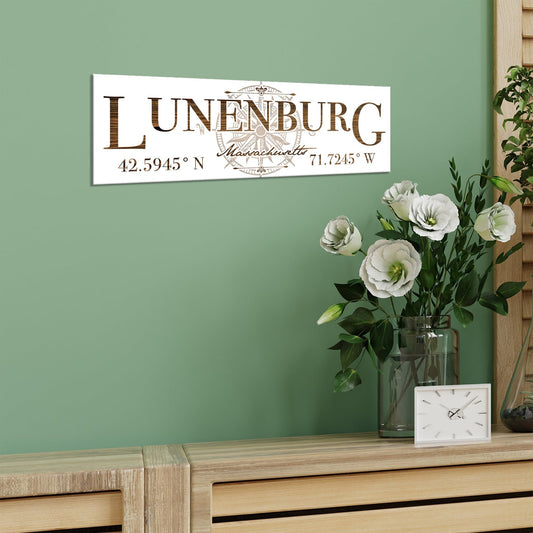 Decorative sign with 'Lunenburg, Massachusetts' and coordinates on a green wall above a wooden surface with flowers and a clock.