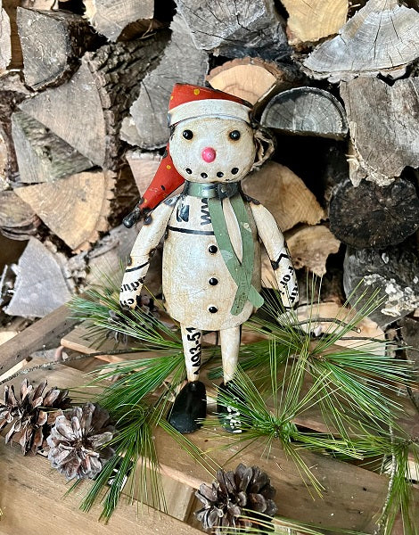 Snowman figurine with a red hat and green scarf standing among stacked firewood and pine branches.