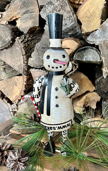 Decorative snowman figurine with a top hat and scarf, standing among stacked firewood and pine branches.
