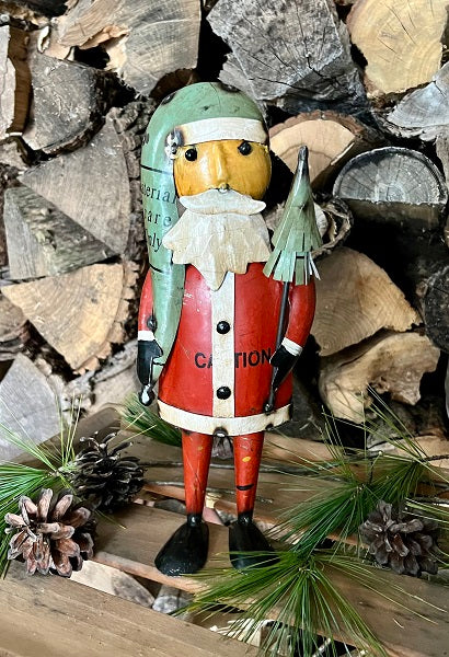 Santa Claus figurine with a green hat and red outfit standing against a background of stacked logs and pine branches.