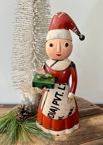 Decorative figure of a Mrs. Claus holding a green gift, with a pine cone and tree branch in the background.