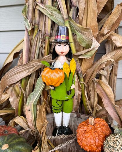 Decorative figurine of a person in a pilgrim hat with pumpkins and corn stalks.