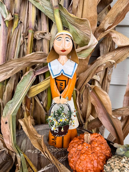 Decorative figurine of a woman holding flowers, surrounded by corn stalks and pumpkins.
