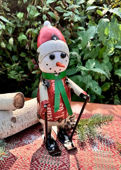 Wooden snowman figurine with a red hat and green scarf, standing on a textured surface with greenery in the background.