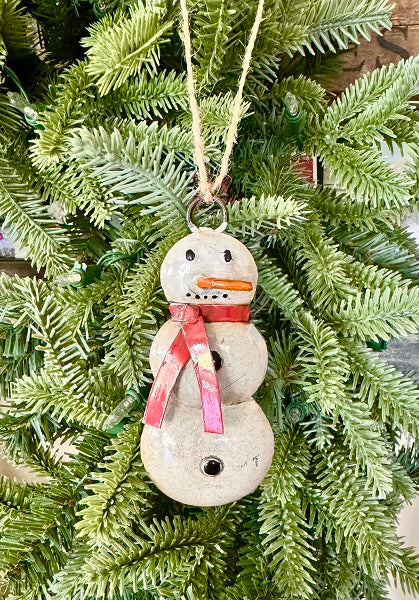 A rustic three-tiered snowman Christmas ornament hanging from a jute string on a green pine tree. The ornament has a weathered beige stone-like finish with a pink and red striped scarf, a small orange carrot nose, and black button eyes.