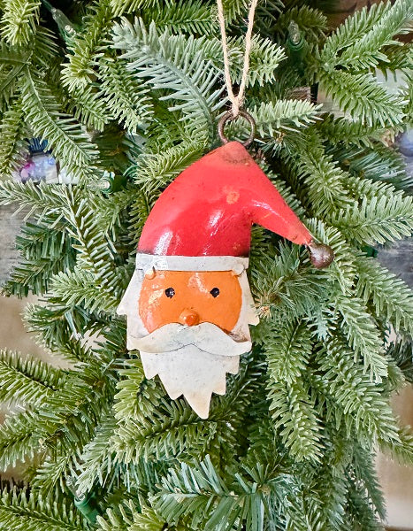 A rustic metal Santa Claus Christmas ornament hanging from a green pine tree branch. The ornament features a red hat, an orange face, a white beard, and a small metal bell at the tip of the hat.