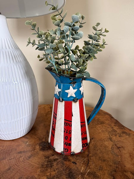 Decorative pitcher with American flag design holding greenery on a wooden surface.